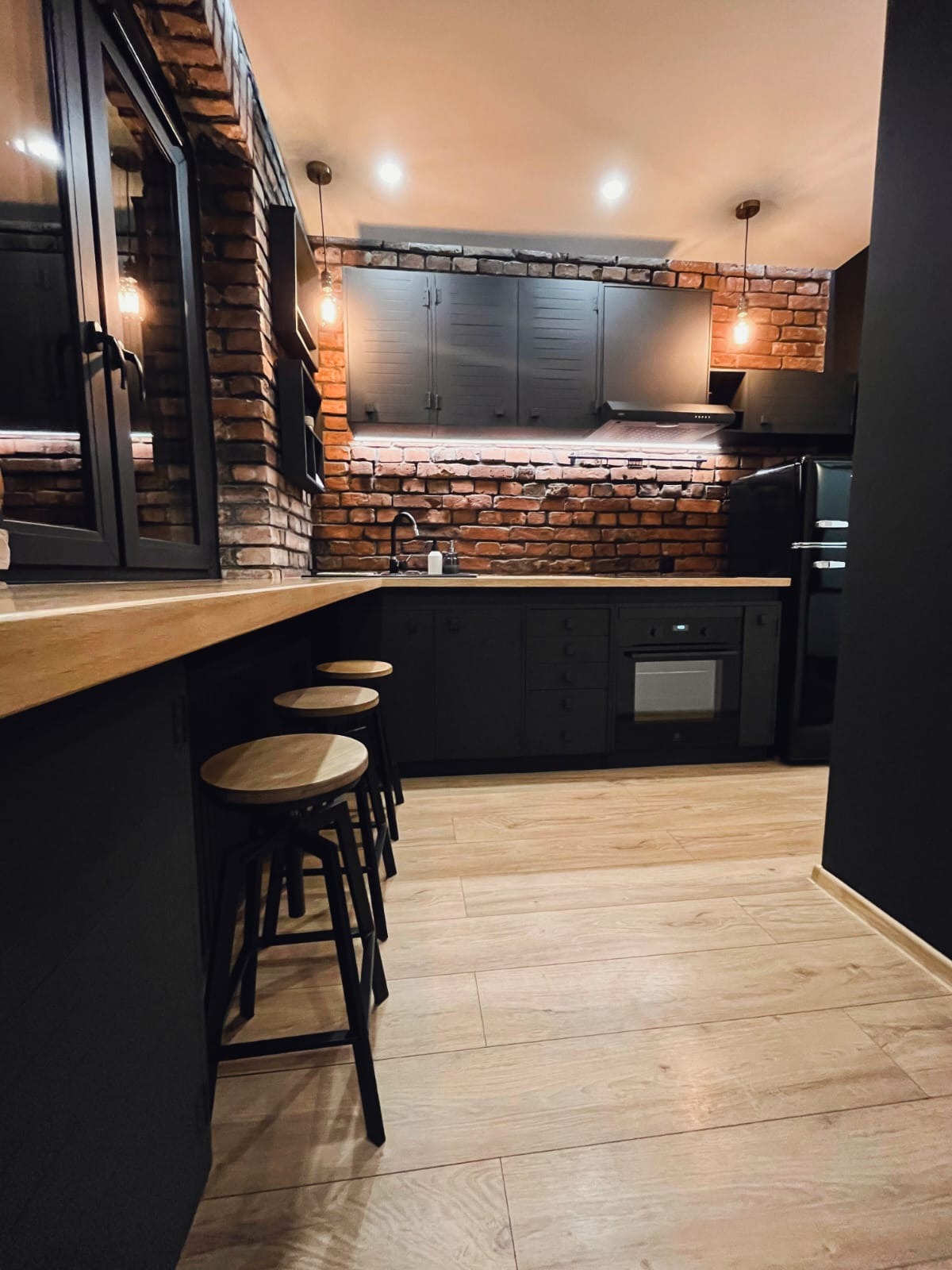 Loftowa kuchnia z ceglaną ścianą, czarnymi szafkami i drewnianym blatem. Trzy hokery przy wyspie. Oświetlenie punktowe i drewniana podłoga.