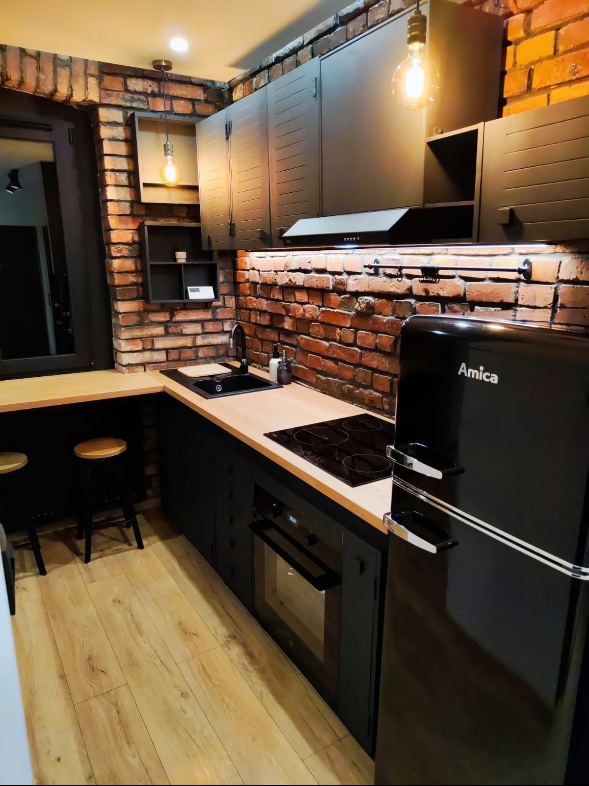 Loftowa kuchnia z ceglaną ścianą, czarnymi szafkami i drewnianym blatem. Widoczny czarny zlew, płyta indukcyjna i lodówka Amica w stylu retro.