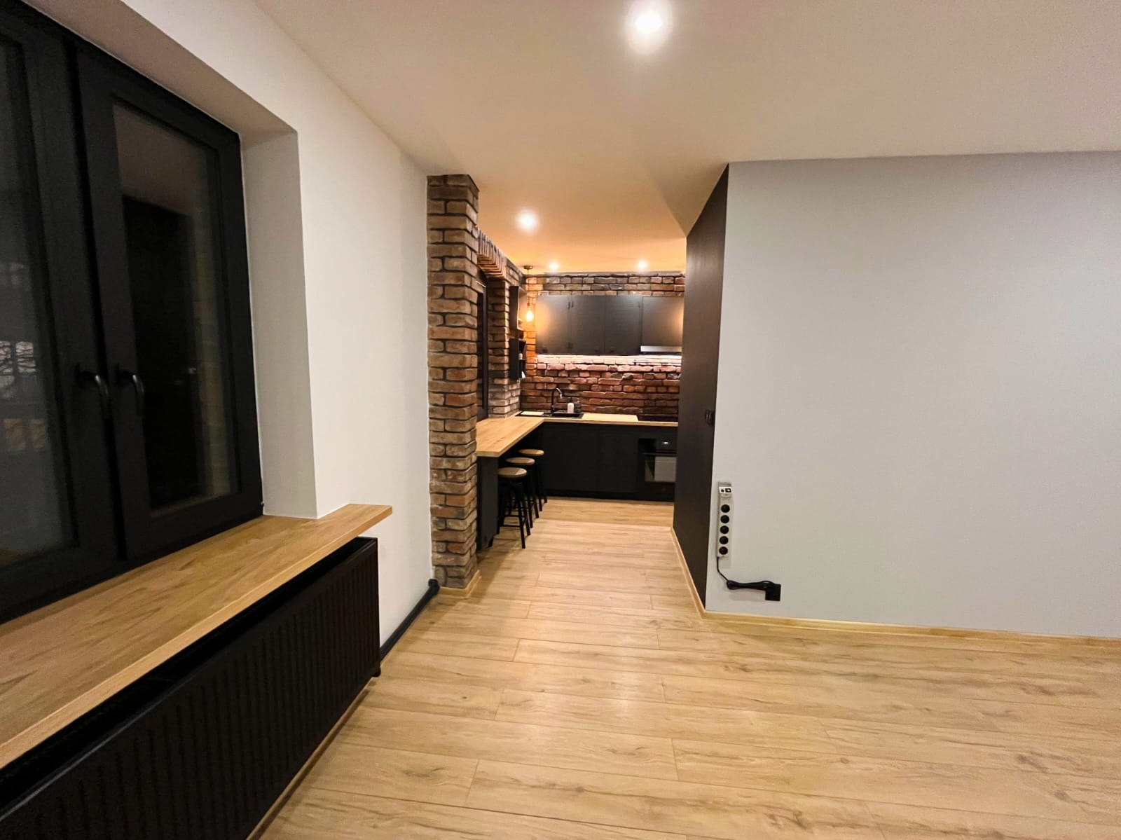 Loftowe wnętrze po remoncie: drewniana podłoga, ceglana ściana w kuchni, czarne meble i okna. Widok na odnowione pomieszczenie z perspektywy wejścia.
