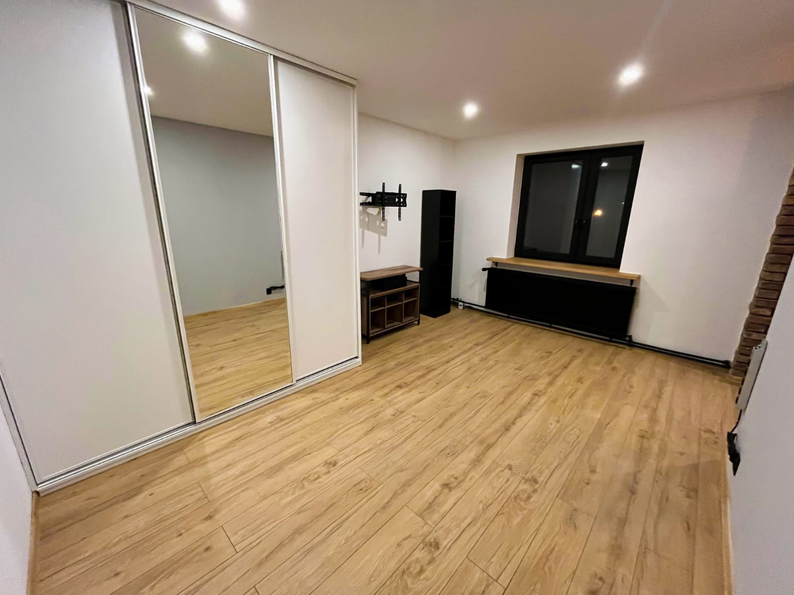 Jasne panele podłogowe w pokoju z szafą przesuwną z lustrem, czarnym grzejnikiem pod oknem i półkami. Widoczny fragment ściany z cegły.