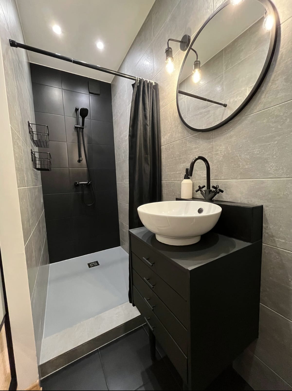 Nowoczesna łazienka w szaro-czarnej tonacji: kabina prysznicowa z czarnymi płytkami, umywalka nablatowa na czarnej szafce, okrągłe lustro z oświetleniem.