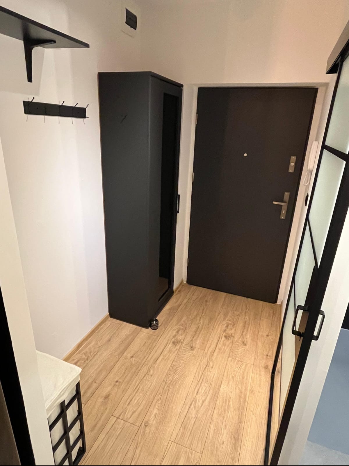 Nowoczesny przedpokój z drewnianą podłogą, czarną szafką na kółkach i drzwiami. Minimalistyczny design, oświetlenie górne. Widok na wnętrze mieszkania.