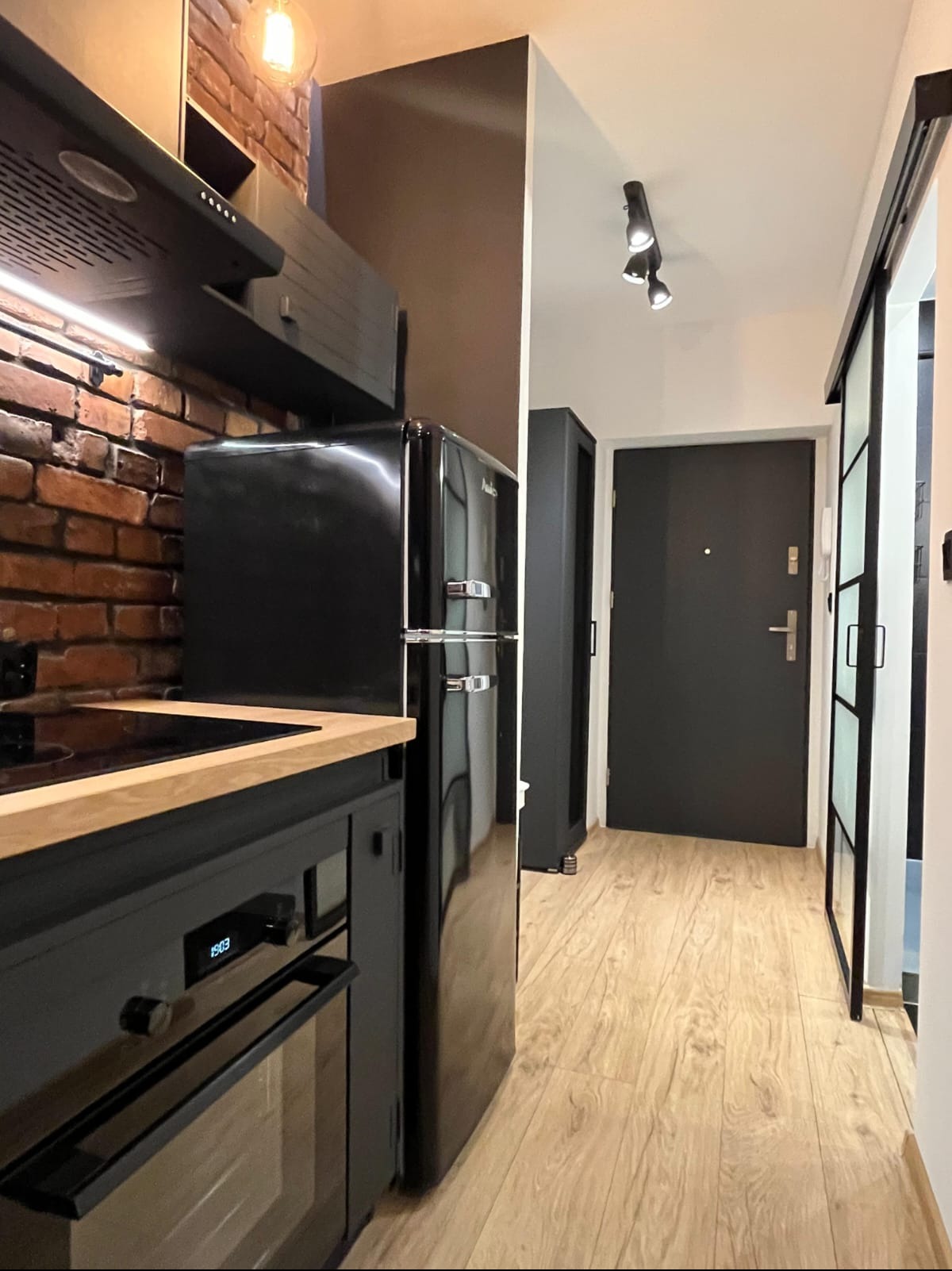 Czarne meble kuchenne z drewnianym blatem i retro lodówką Amica na tle ceglanej ściany. Widok na przedpokój z drewnianą podłogą i czarnymi drzwiami.
