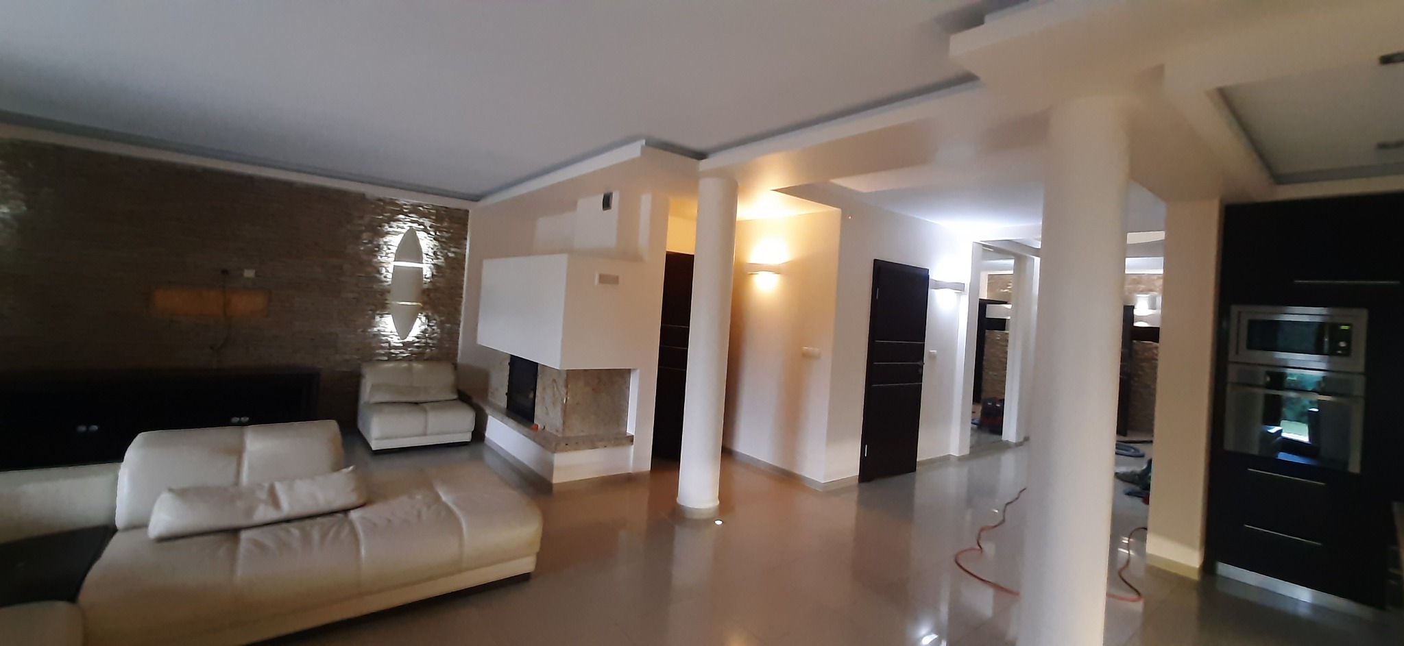 Przestronny salon po renowacji z beżowymi ścianami, kominkiem obłożonym kamieniem i błyszczącą podłogą. Widoczne kolumny i nowoczesne oświetlenie.