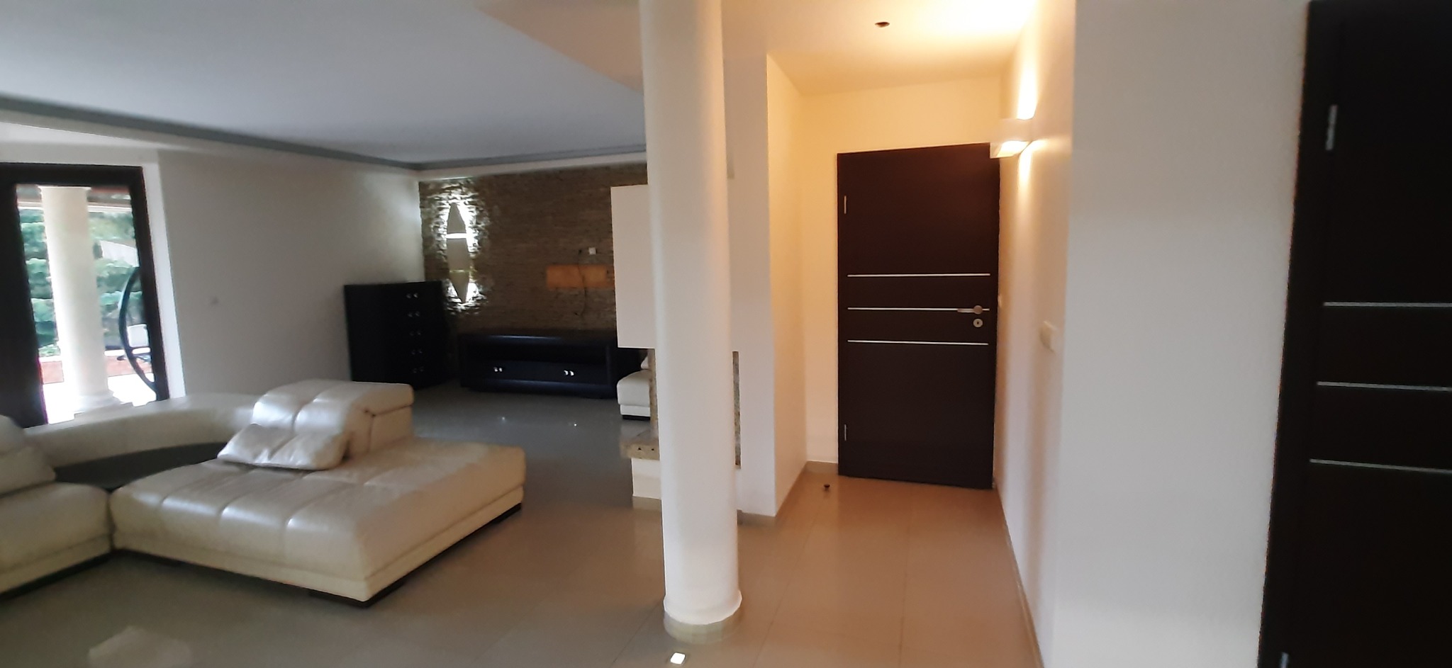 Jasne wnętrze domu z beżową sofą, kolumnami i dekoracyjną ścianą z kamienia. Widoczna ciemna szafka i drzwi. Oświetlenie punktowe.