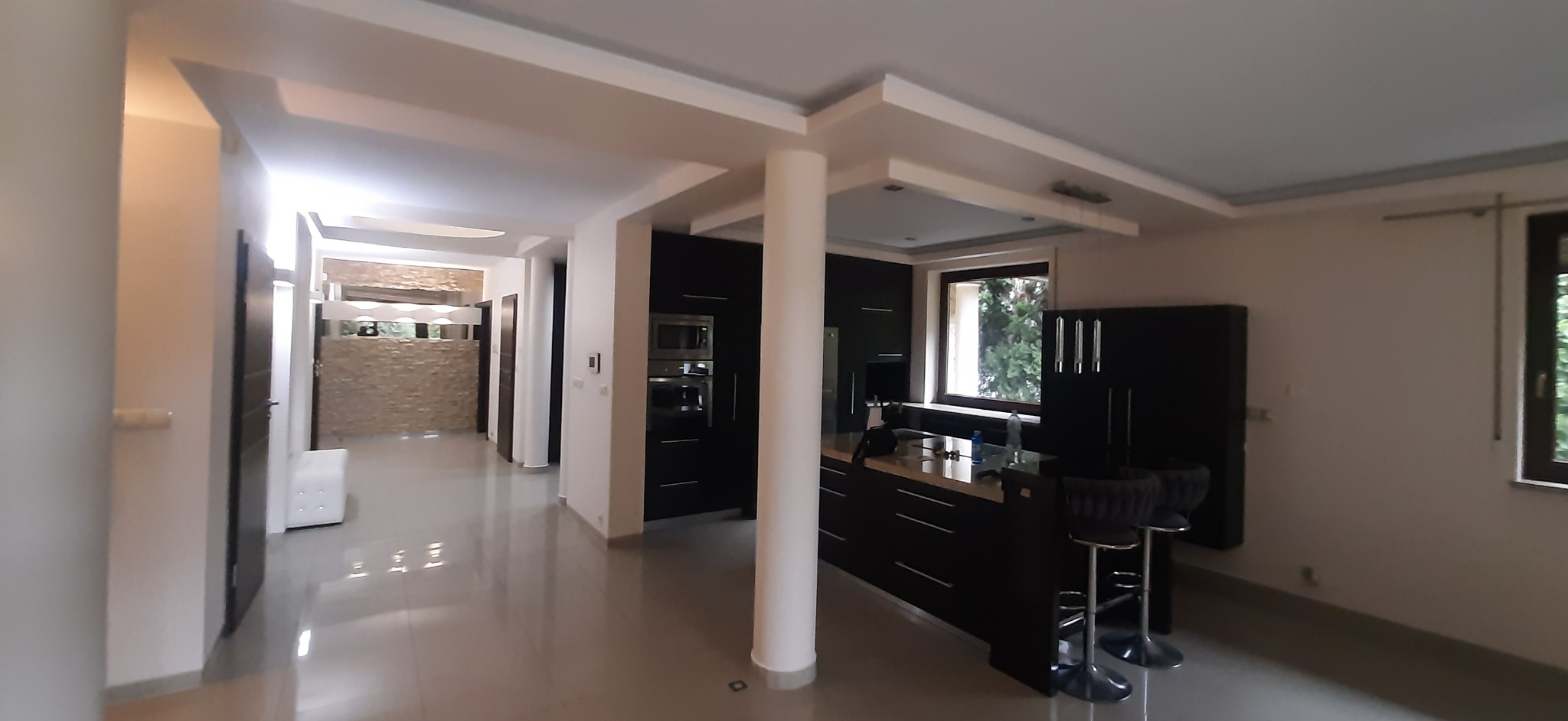 Nowoczesna kuchnia z wyspą i barkiem, czarne meble, jasne ściany, kolumna podtrzymująca sufit, widok na salon z dekoracyjną ścianą z kamienia.