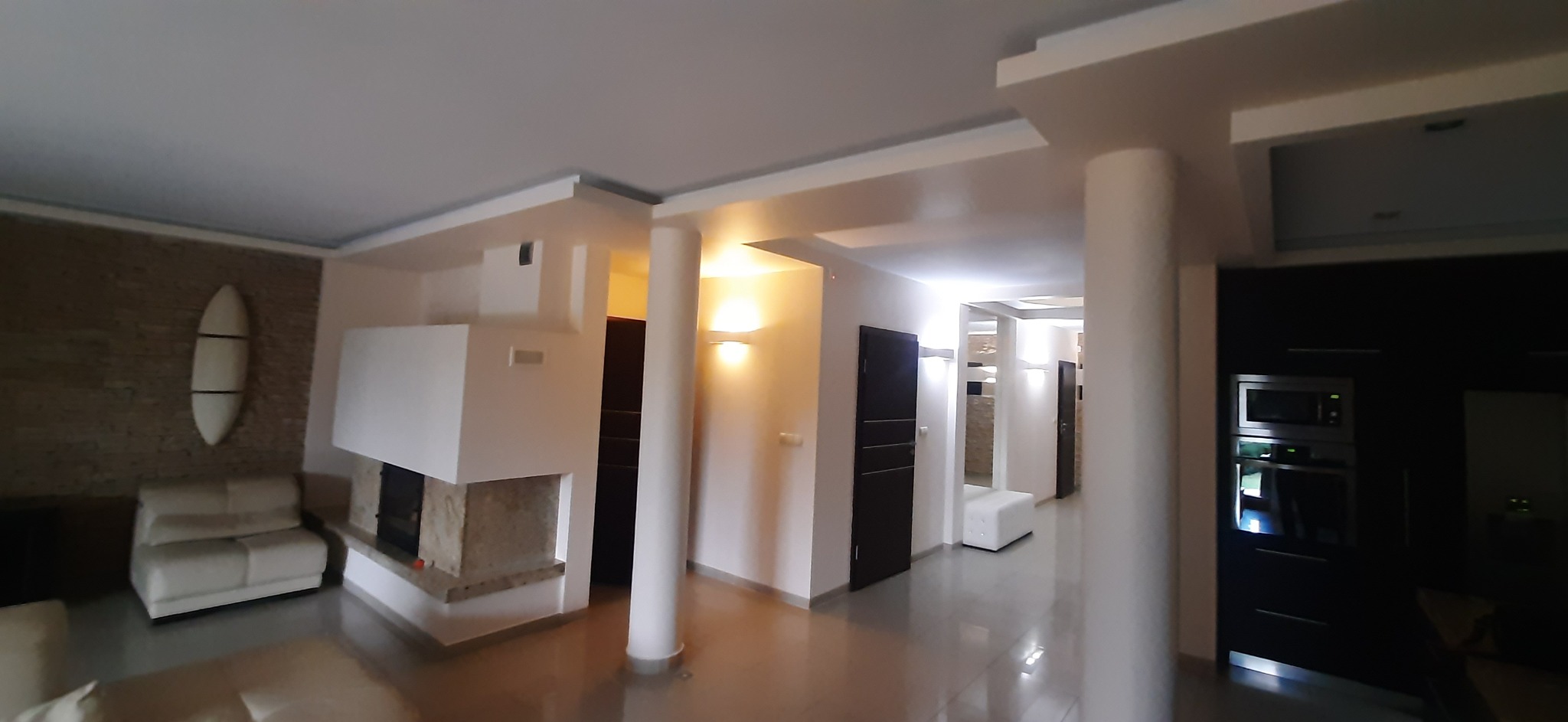 Przestronne wnętrze salonu z kominkiem i elementami dekoracyjnymi, utrzymane w jasnej kolorystyce z akcentami kamienia i drewna. Widoczne kolumny i nowoczesne oświetlenie.