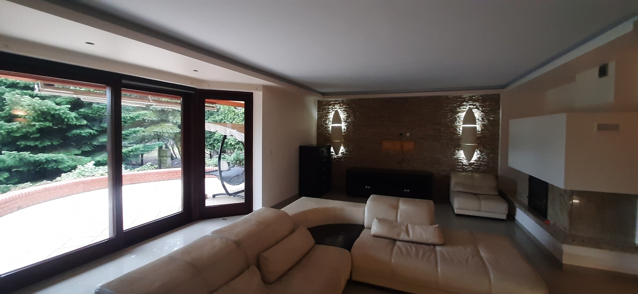 Jasne wnętrze salonu z dużym oknem na ogród, beżowa sofa, ściana z kamienia dekoracyjnego z oświetleniem. Nowoczesny design, przestronność i naturalne światło.