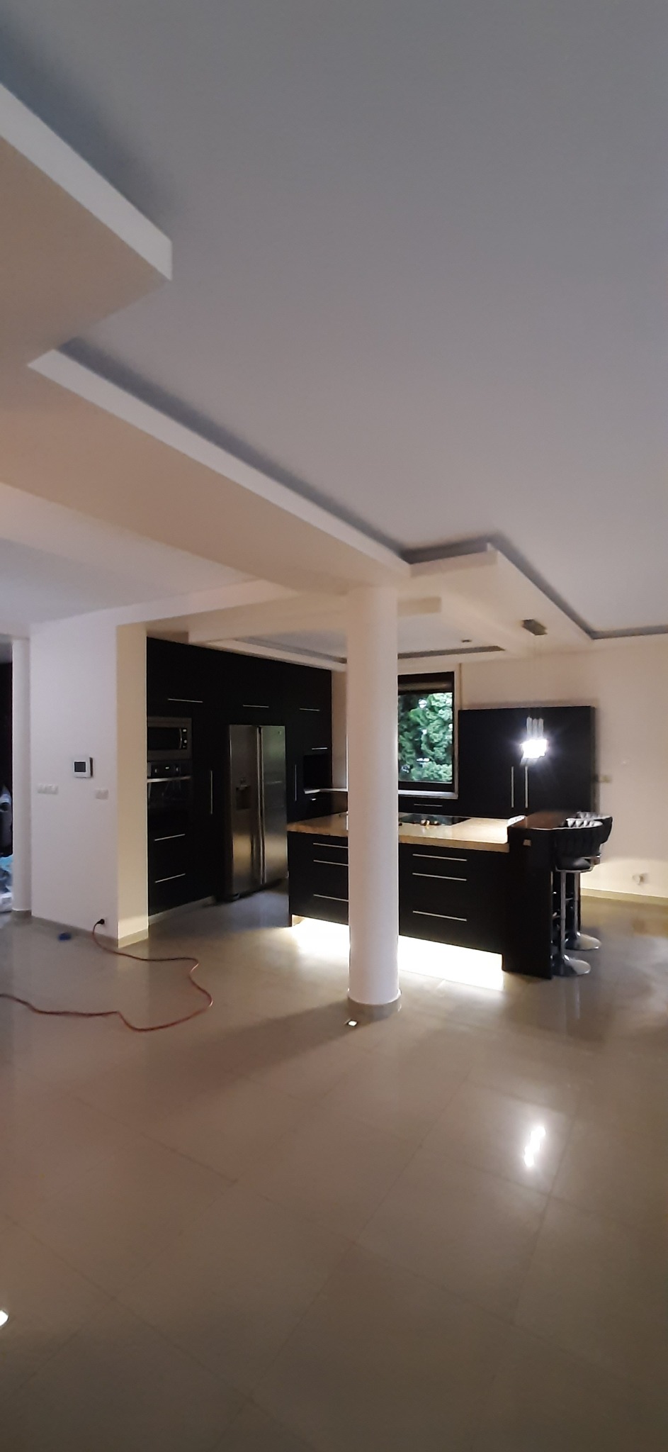 Nowoczesne wnętrze: czarna zabudowa kuchenna z wyspą i hokerami, jasna podłoga i sufit z podświetleniem. Widok na okno z zielenią. Minimalistyczny design.