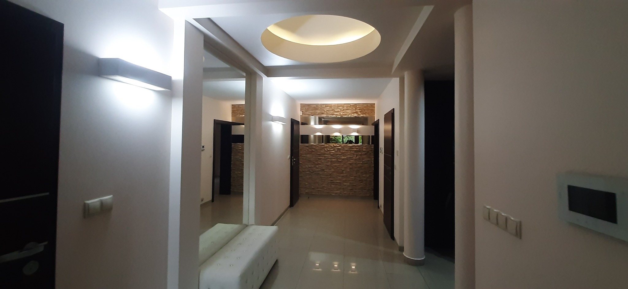Nowoczesny korytarz z oświetleniem LED, dekoracyjną ścianą z kamienia i okrągłym plafonem. Biała ławka z pikowaniem dodaje elegancji. Widok na wnętrze mieszkania.