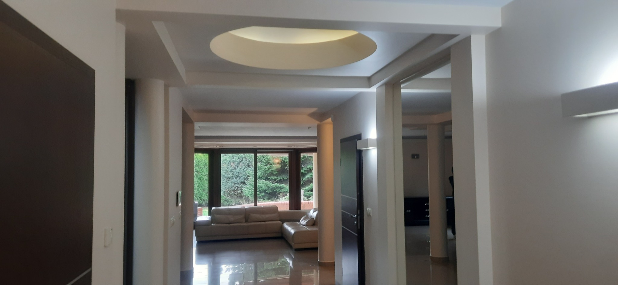 Nowoczesne wykończenie wnętrza: białe ściany, kolumny i sufit z okrągłym oświetleniem. Widok na salon z dużym oknem i sofą. Błyszcząca podłoga.