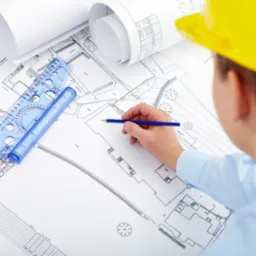 Osoba w kasku nanosi poprawki na plan architektoniczny z użyciem ołówka i linijki. Widoczne zwinięte plany i szczegóły projektu.