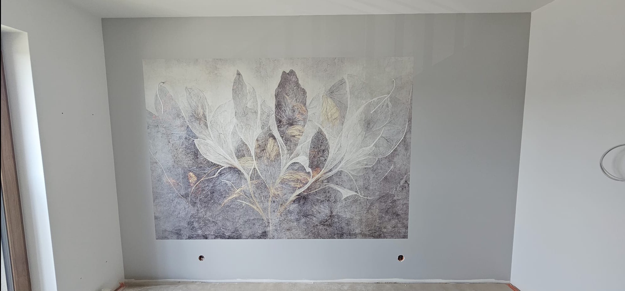 Ściana w szarym odcieniu z dużym, abstrakcyjnym motywem roślinnym. Widoczne otwory w ścianie, prawdopodobnie pod gniazdka elektryczne. Wnętrze w trakcie wykończenia.