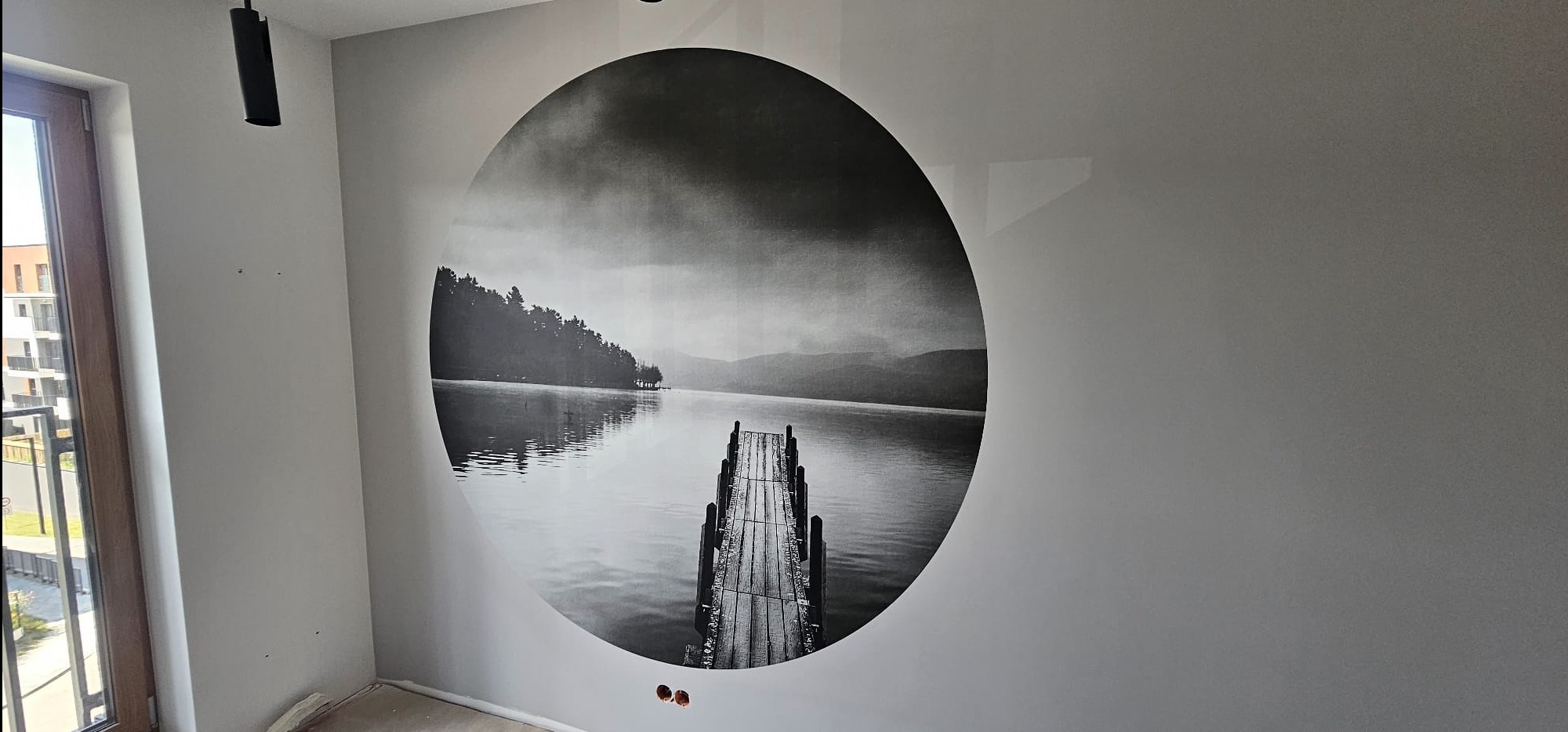 Ściana z okrągłym, czarno-białym fototapetą przedstawiającą jezioro i pomost. Widoczne gniazdka elektryczne i fragment okna. Minimalistyczne wykończenie wnętrza.