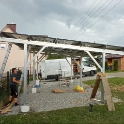 Usługi hydrauliczno-budowlane Adrian Bugała - Montaż paneli solarnych na aluminiowej konstrukcji w ogrodzie. Widoczny monter z drabiną, biały bus i dwa domy w tle. Pochmurne niebo.