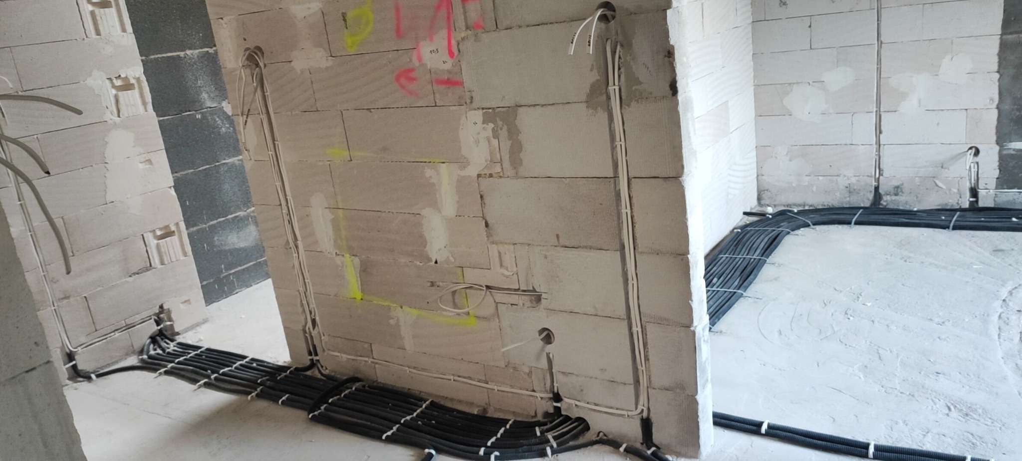 Instalacja elektryczna w surowym wnętrzu: kable w peszlach ułożone wzdłuż ścian z betonu komórkowego, pionowe przewody w bruzdach. Widoczne oznaczenia na ścianach.