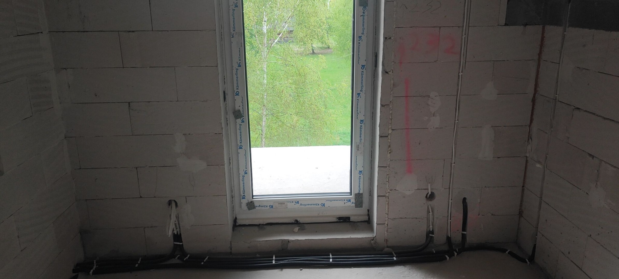 Surowy stan: instalacja elektryczna w ścianach z betonu komórkowego. Widoczne przewody prowadzone wzdłuż ściany pod oknem i pionowo w górę. Otwór okienny z widokiem na zieleń.