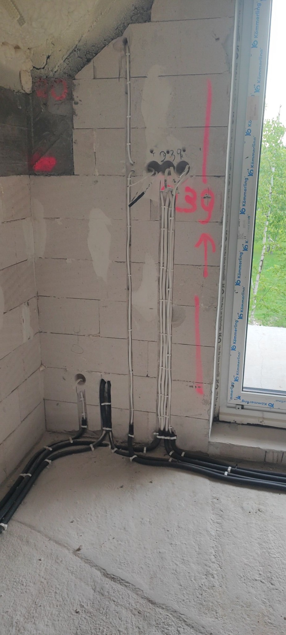 Wnętrze w trakcie prac: białe przewody elektryczne prowadzone wzdłuż ściany z betonu komórkowego, czarne rury instalacyjne przy podłodze. Widoczne oznaczenia na ścianie.