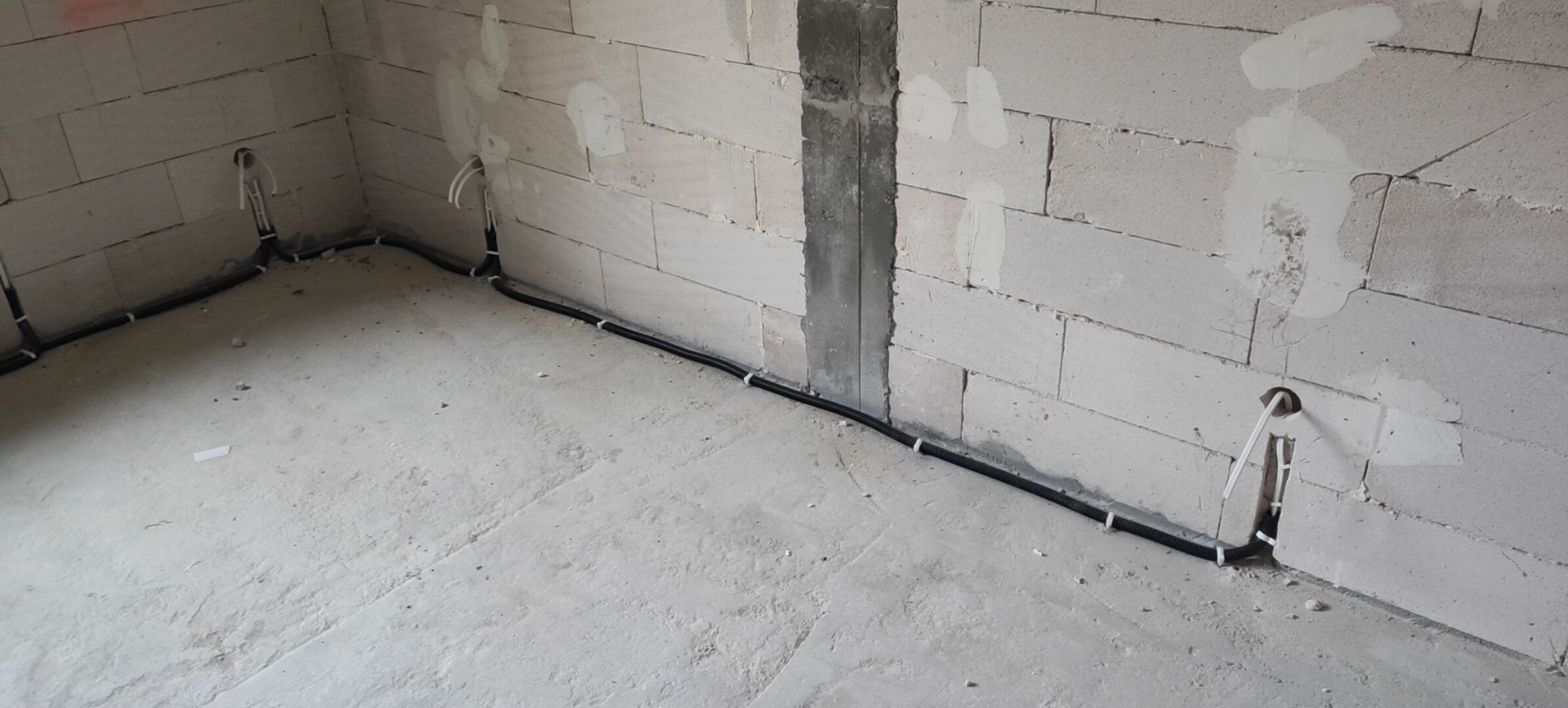 Wnętrze w trakcie remontu: widoczne kable elektryczne w peszlu, prowadzone wzdłuż ściany z betonu komórkowego, przytwierdzone białymi uchwytami. Otwory w ścianach przygotowane pod gniazdka.