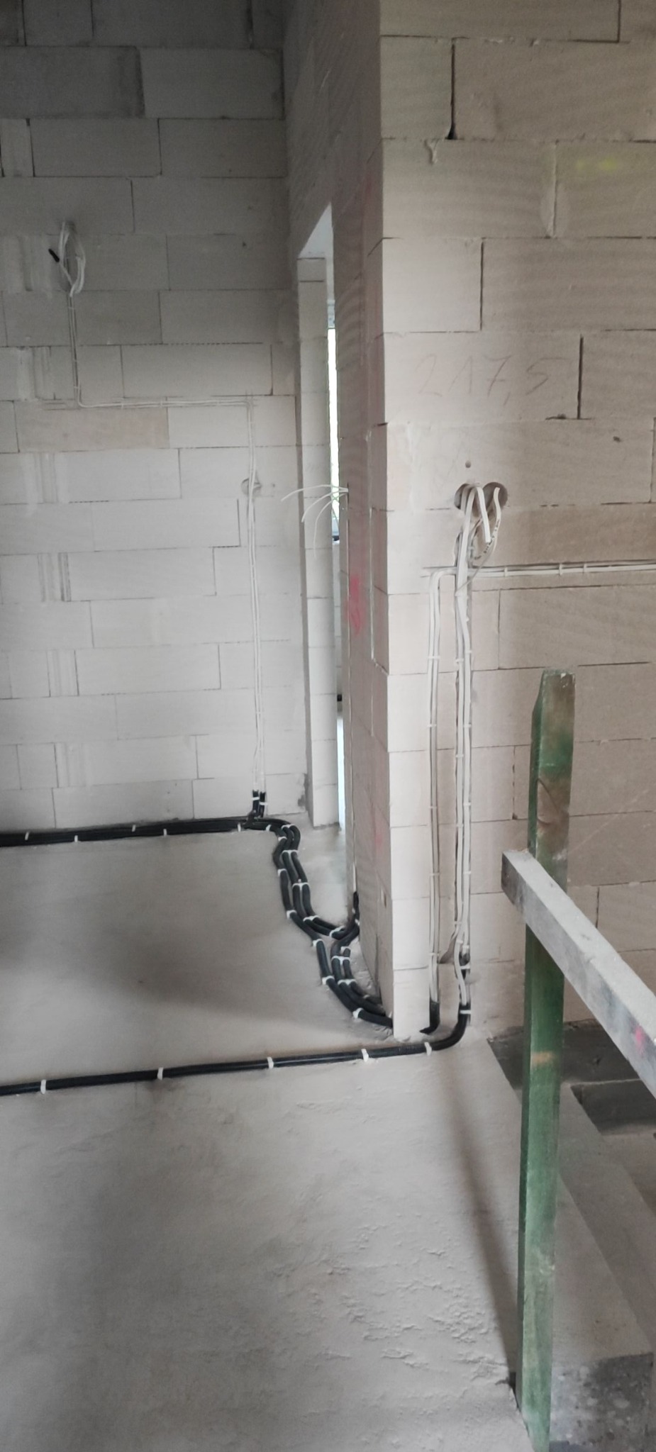 Surowe ściany z betonu komórkowego i widoczne, czarne rury instalacji elektrycznej ułożone wzdłuż podłogi, białe kable wystające ze ściany.