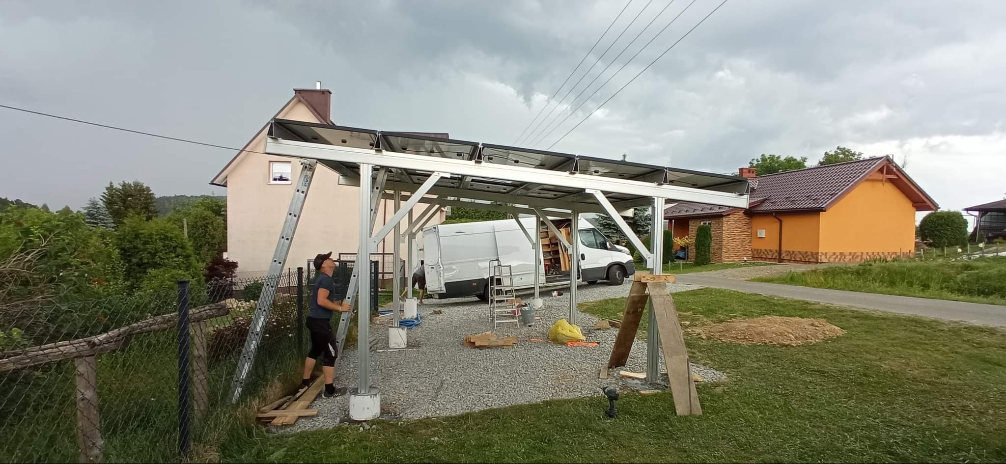 Montaż paneli solarnych na aluminiowej konstrukcji w ogrodzie. Widoczny monter z drabiną, biały bus i dwa domy w tle. Pochmurne niebo.