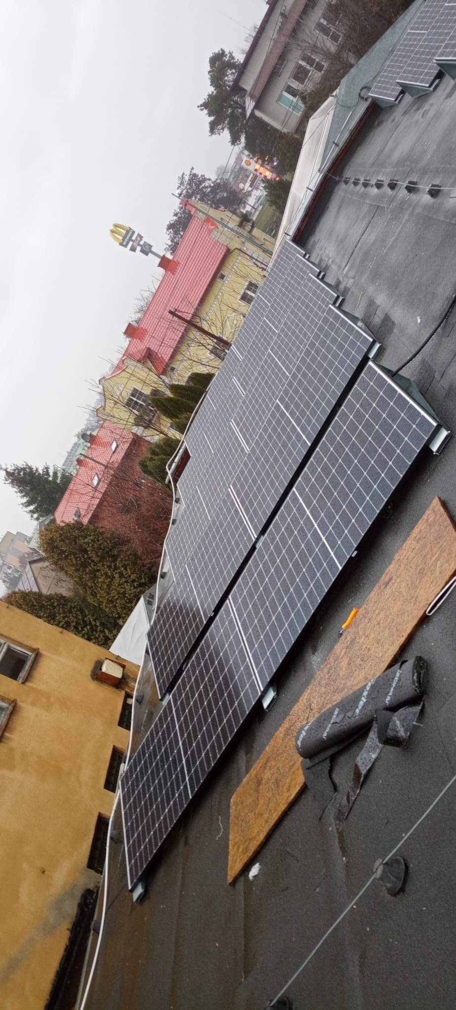 Montaż paneli słonecznych na dachu budynku z widocznym fragmentem miasta w tle, w tym logo McDonald's. Panele ułożone rzędami, widoczne materiały montażowe.