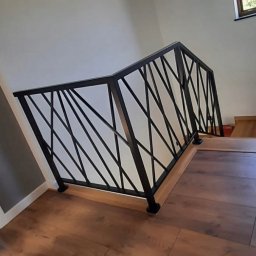 ProSteel - Nowoczesna, czarna balustrada schodowa z geometrycznym wzorem, zamontowana na drewnianych schodach w jasnym wnętrzu. Widok z dołu, podkreślający unikalny design.