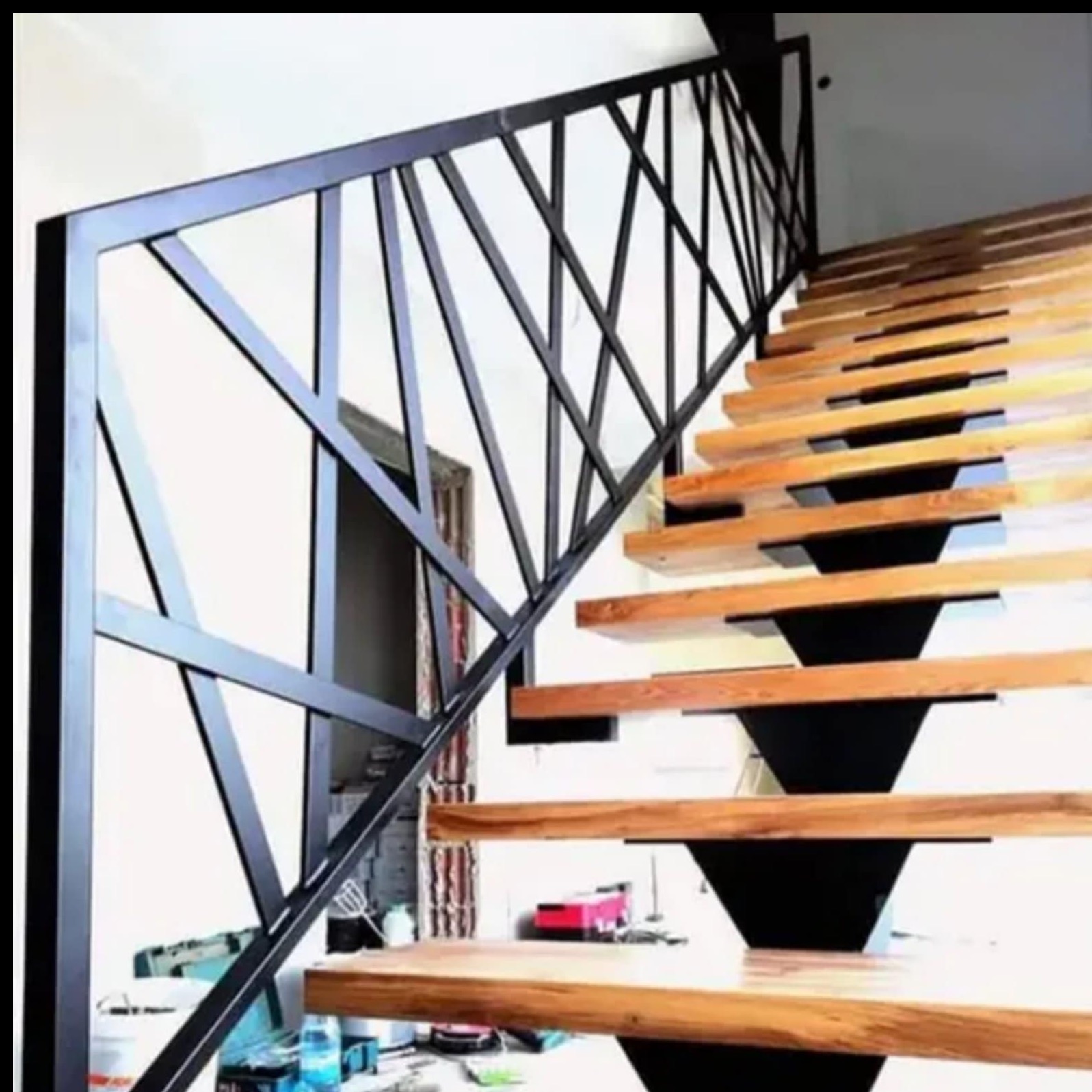 Nowoczesne schody z drewnianymi stopniami i geometryczną, czarną metalową balustradą. Widok z dołu, częściowo widoczne akcesoria montażowe.