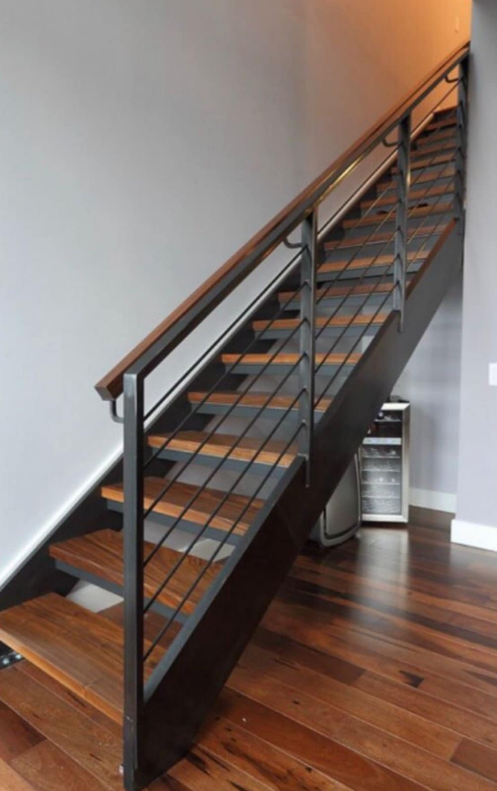 Nowoczesne schody z drewnianymi stopniami i metalową balustradą z poziomymi prętami, widoczne z dołu, oświetlone ciepłym światłem.
