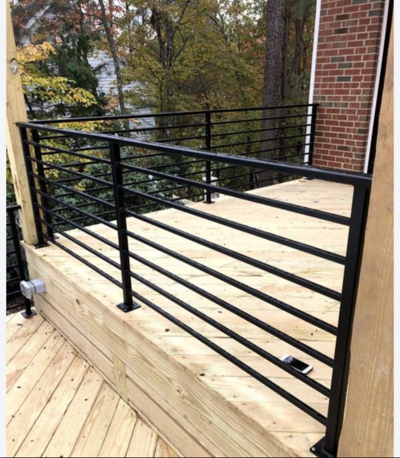 Nowoczesna, czarna balustrada z poziomych prętów na drewnianym tarasie. Widoczny smartfon leżący na podłodze tarasu. W tle drzewa i ściana budynku.