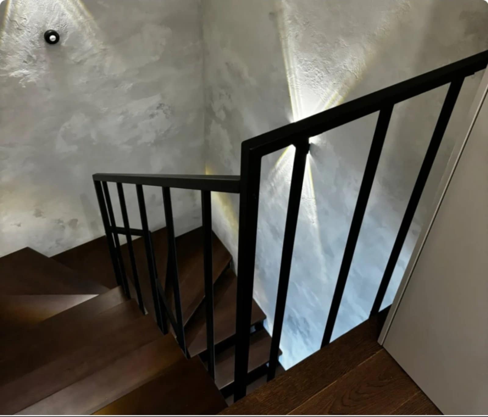 Nowoczesne, drewniane schody z czarną, prostą balustradą. Ściana oświetlona światłem punktowym, z efektem dekoracyjnego tynku. Ujęcie z góry, podkreślające geometrię.