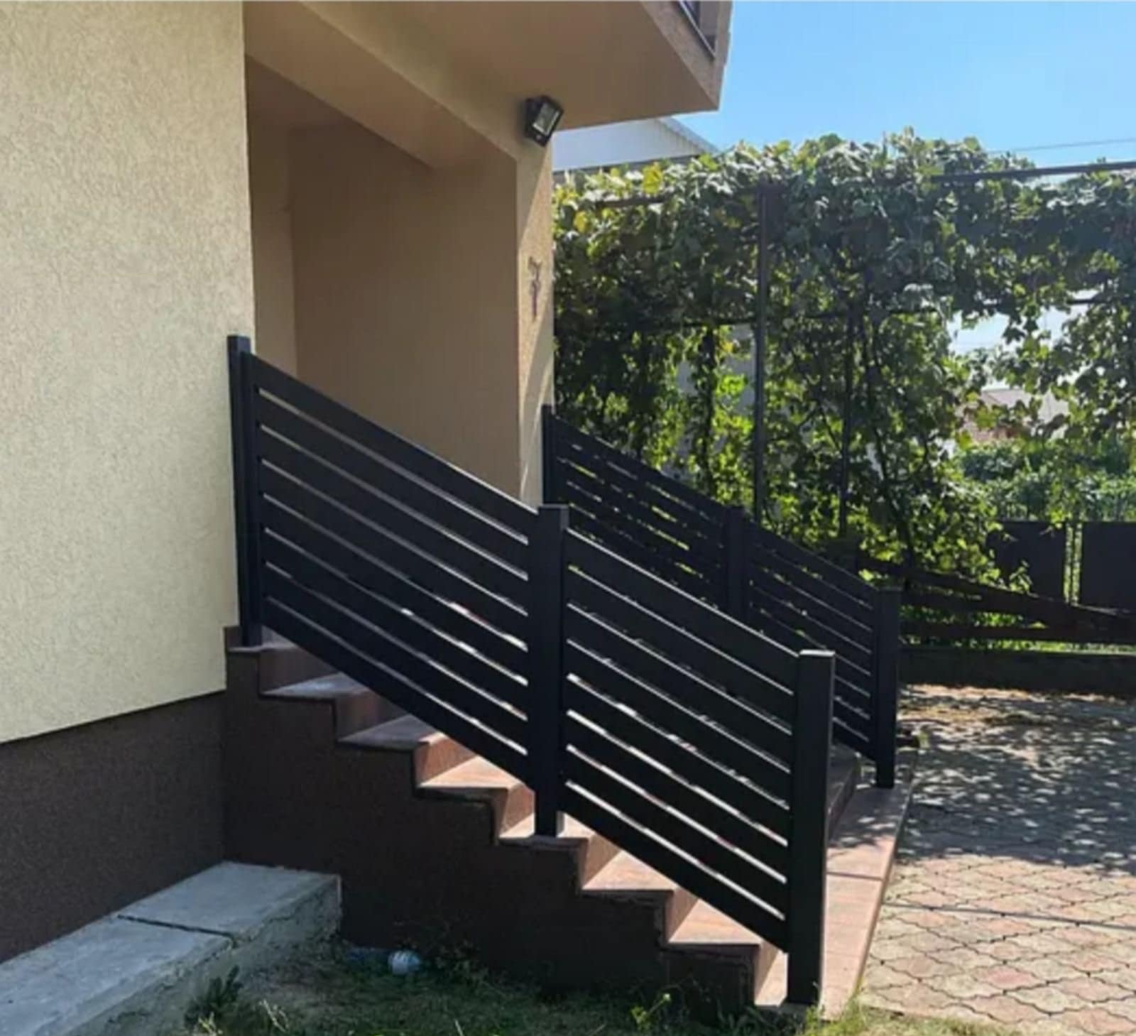 Czarne, poziome panele balustrady schodowej z metalu, prowadzące do wejścia budynku z beżową elewacją i zielonymi roślinami w tle.