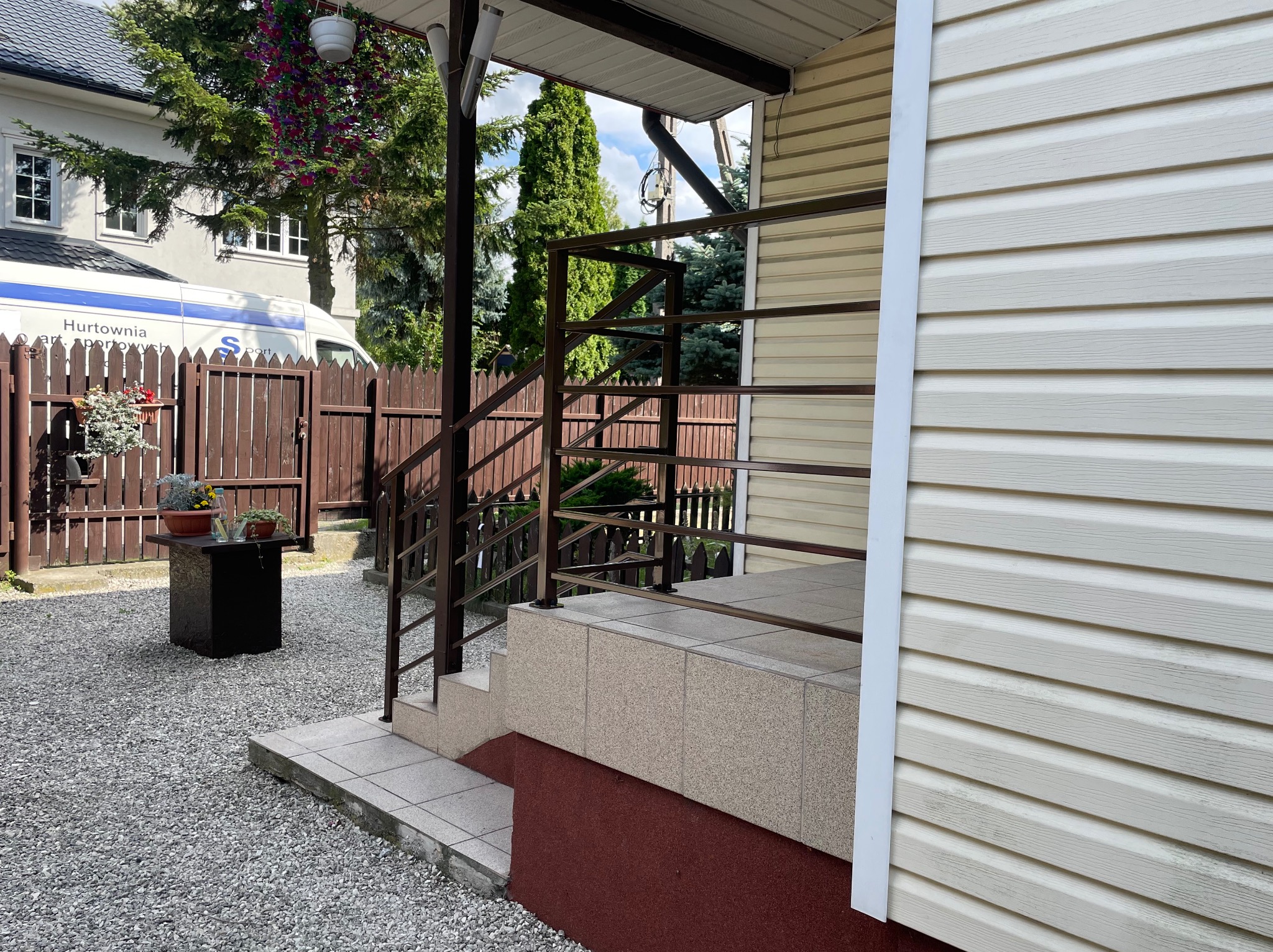 Metalowa balustrada schodowa w kolorze brązowym, prowadząca do wejścia domu z sidingiem. Widok z boku, na pierwszym planie szara nawierzchnia z kamyków.