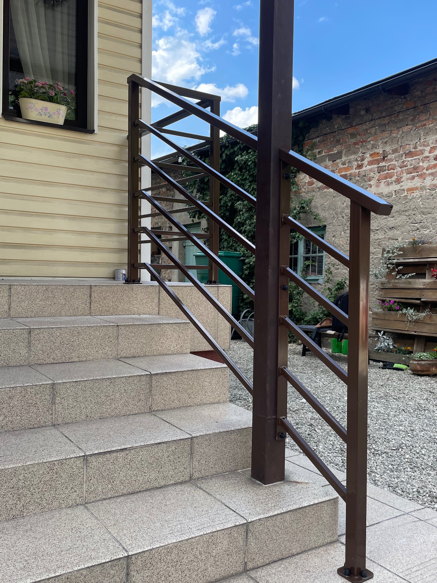 Zewnętrzne schody z brązową metalową balustradą. Widoczne płytki na schodach i elementy mocujące balustradę. W tle budynek z cegły i zieleń.