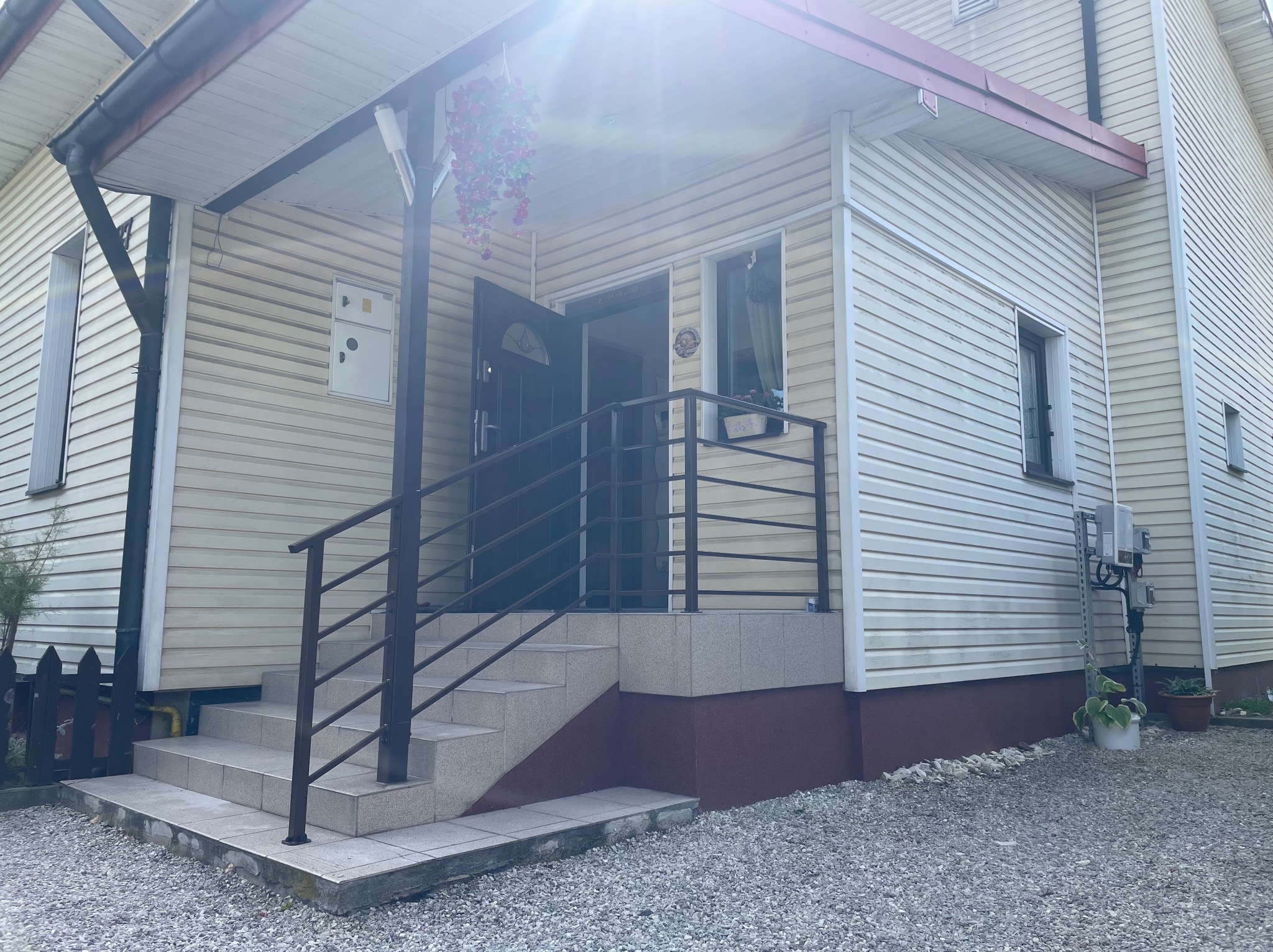 Zewnętrzne schody z ciemną, metalową balustradą prowadzące do wejścia domu z sidingiem. Widoczny fragment ogrodu i słoneczne odbicie na budynku.