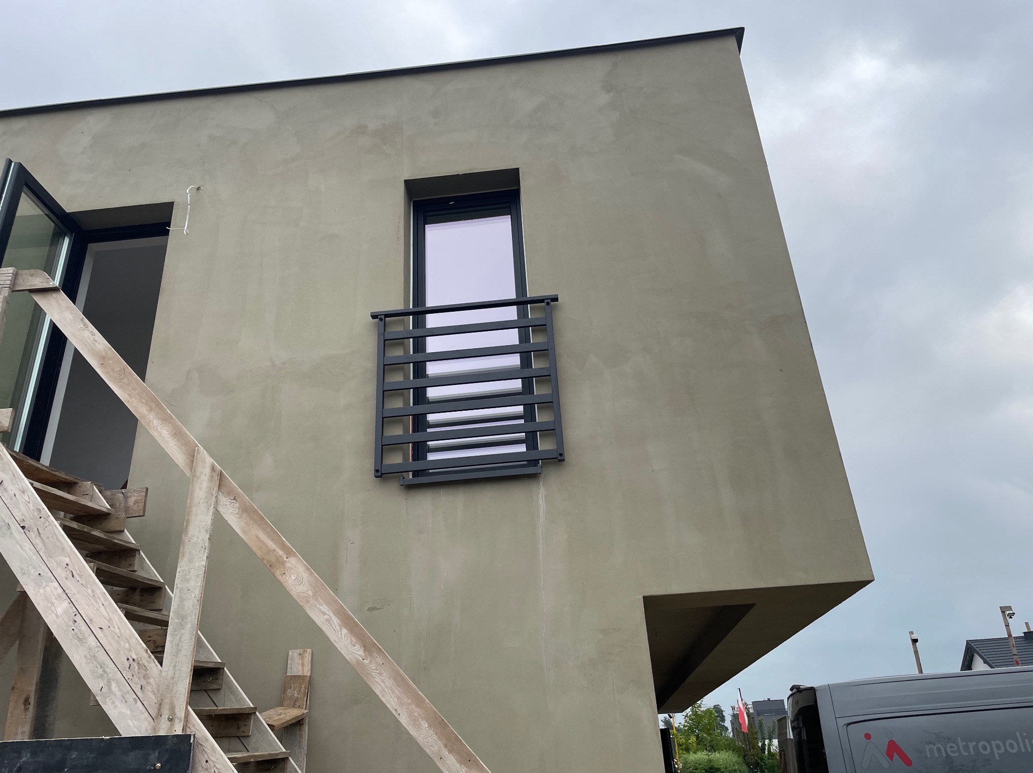 Szara elewacja budynku z oknem zabezpieczonym czarną, poziomą balustradą. Widoczne drewniane schody i fragment samochodu dostawczego.