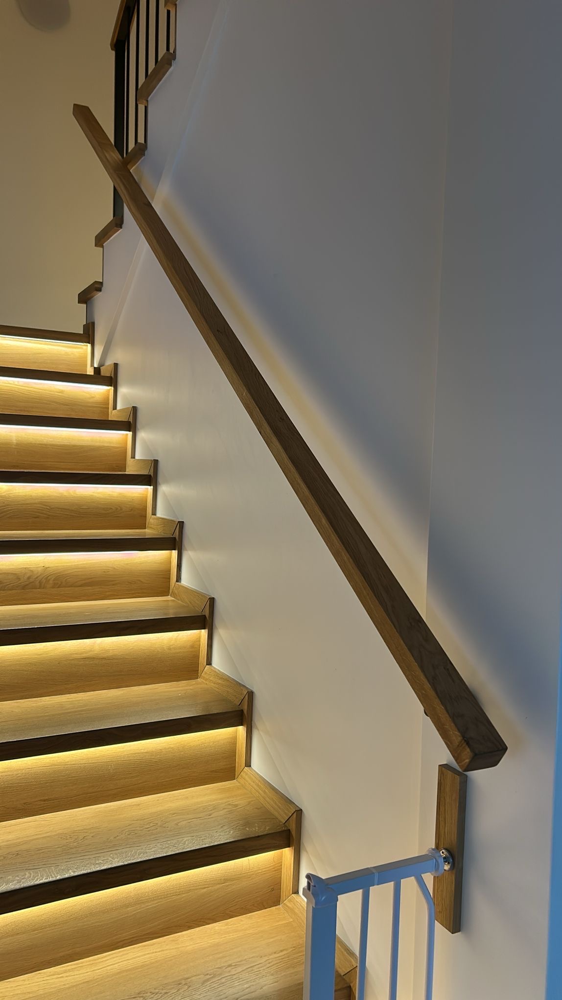 Drewniane schody z podświetleniem LED i drewnianą poręczą, zabezpieczone białą barierką dla dzieci. Nowoczesny design, ciepłe oświetlenie, widok z dołu.