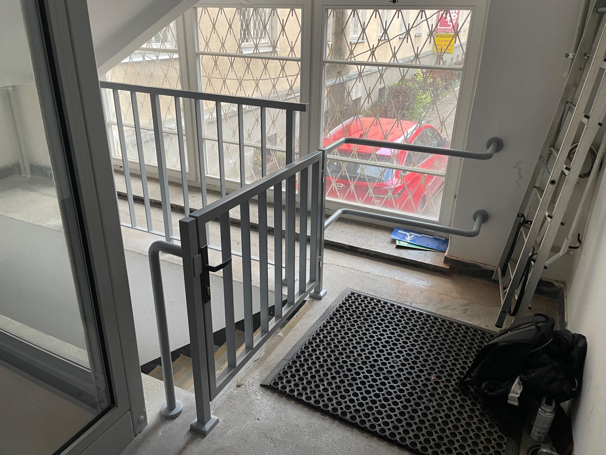 Szara, metalowa barierka schodowa z poręczą, zamontowana przy oknie z kratą. Widoczny fragment schodów, wycieraczka i torba. W tle czerwony samochód.