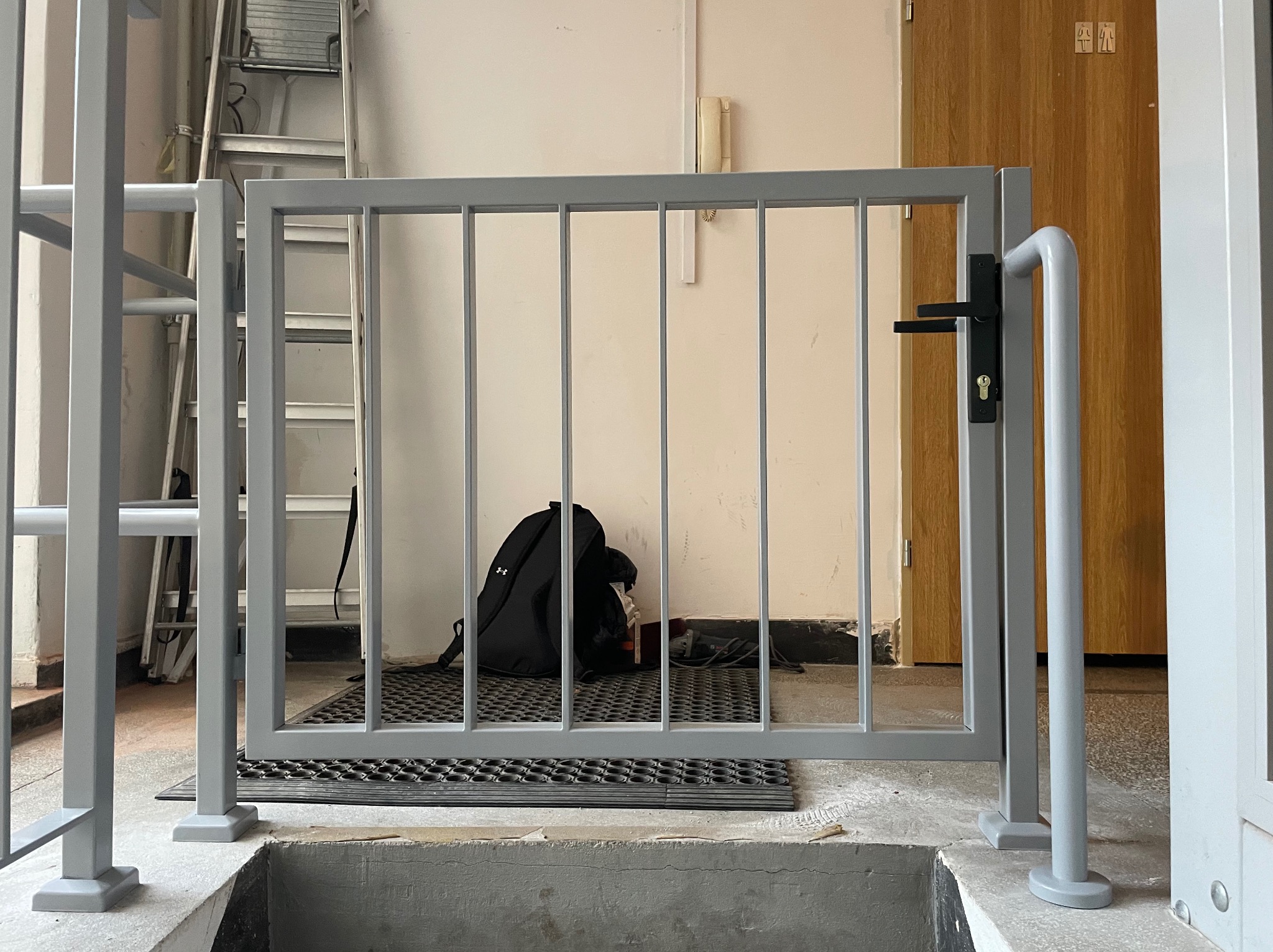 Szara metalowa barierka z zamkiem zabezpieczająca otwór w podłodze, widoczna drabina i torba w tle, wewnątrz budynku.