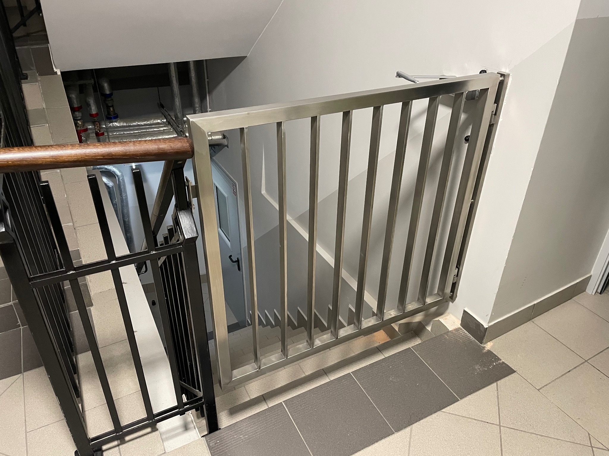 Metalowa barierka schodowa z pionowymi prętami, zamontowana na podeście. Widoczne schody w dół i instalacje. Ujęcie z góry, z bliska, w neutralnym oświetleniu.