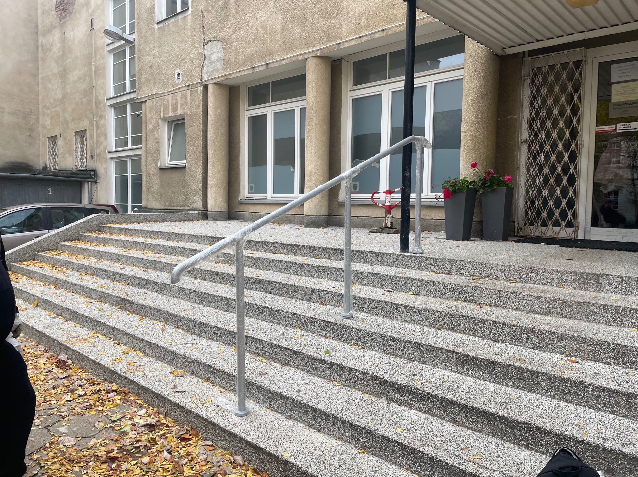Zewnętrzne schody z szarego lastryko z nową, srebrną poręczą zabezpieczoną folią. Jesienne liście na stopniach i chodniku. Budynek w tle.