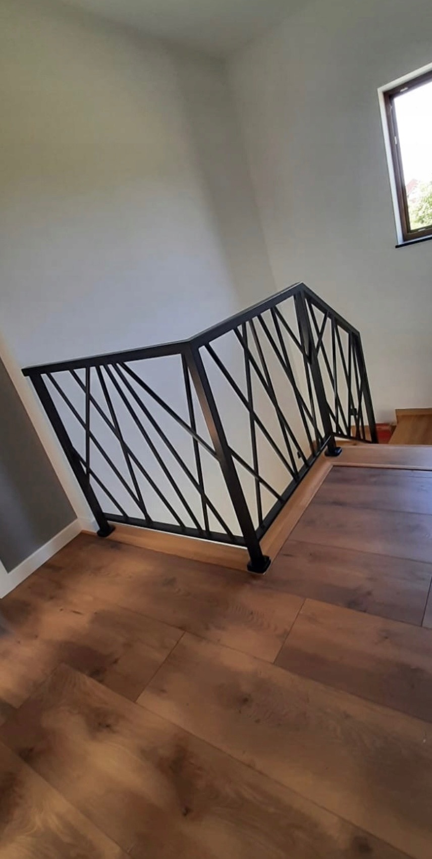 Nowoczesna, czarna balustrada schodowa z geometrycznym wzorem, zamontowana na drewnianych schodach w jasnym wnętrzu. Widok z dołu, podkreślający unikalny design.