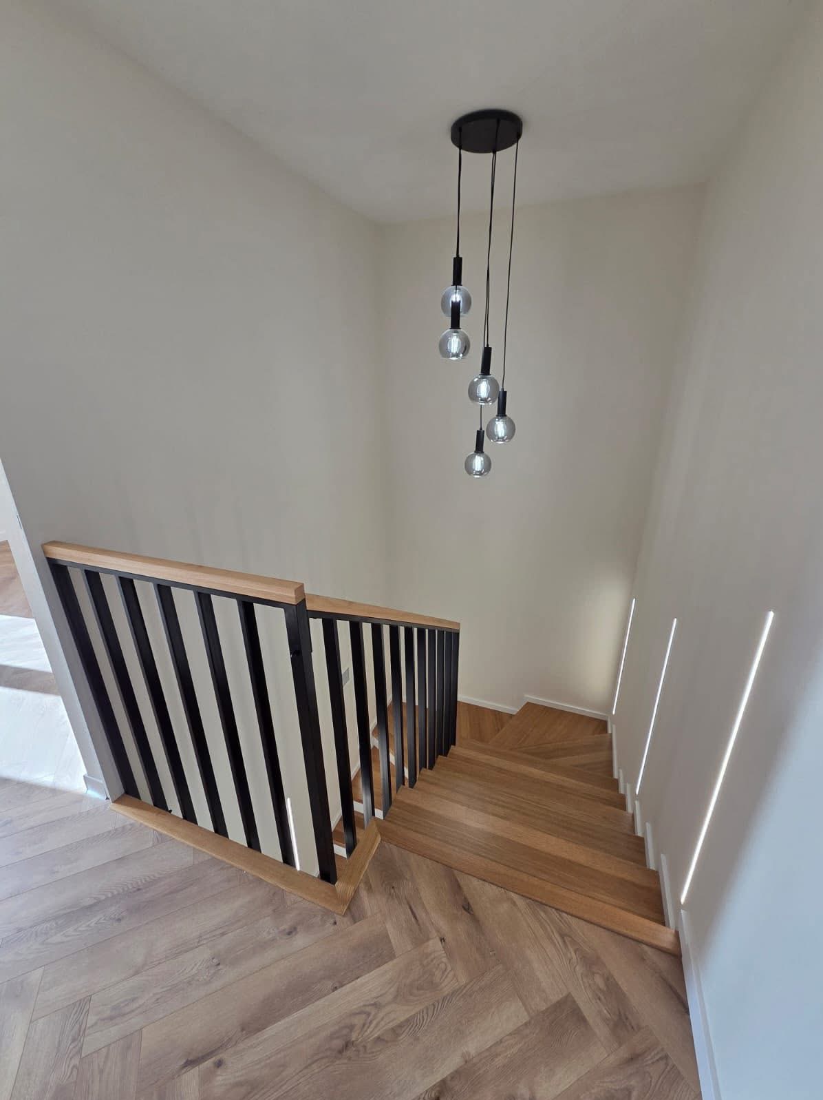 Nowoczesne schody drewniane z czarną balustradą i drewnianym poręczem, oświetlone wiszącą lampą z kilkoma szklanymi kloszami. Widok z góry na dół.