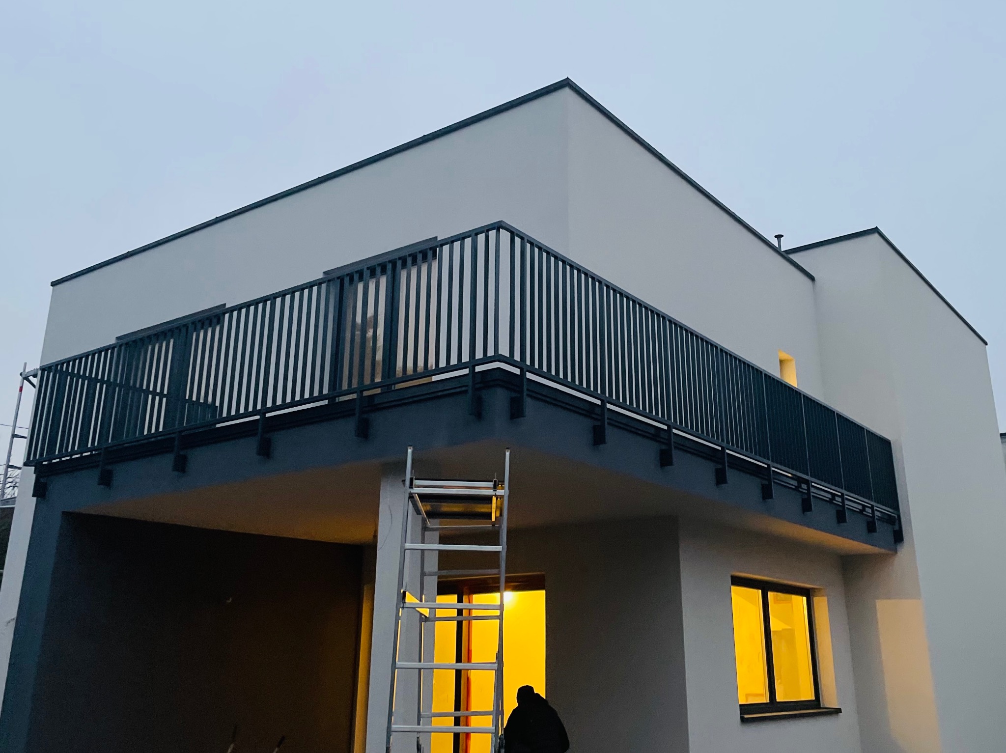 Nowoczesny balkon z czarną, metalową balustradą pionową, widok z dołu na tle szarego budynku. W tle drabina i sylwetka osoby w oświetlonym pomieszczeniu.