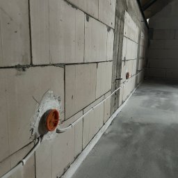 Elmat - Wewnętrzna instalacja elektryczna w surowym budynku: białe kable prowadzone wzdłuż ściany z pustaków, pomarańczowe puszki pod gniazdka. Szara podłoga.