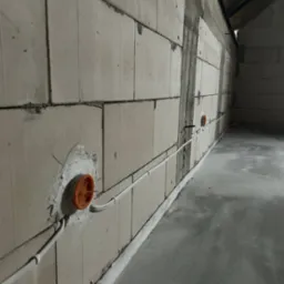 Wewnętrzna instalacja elektryczna w surowym budynku: białe kable prowadzone wzdłuż ściany z pustaków, pomarańczowe puszki pod gniazdka. Szara podłoga.