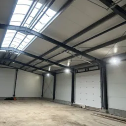 Wnętrze hali magazynowej z oświetleniem LED, widoczne stalowe konstrukcje, białe ściany z paneli i brama segmentowa. Na suficie świetliki doświetlające wnętrze.