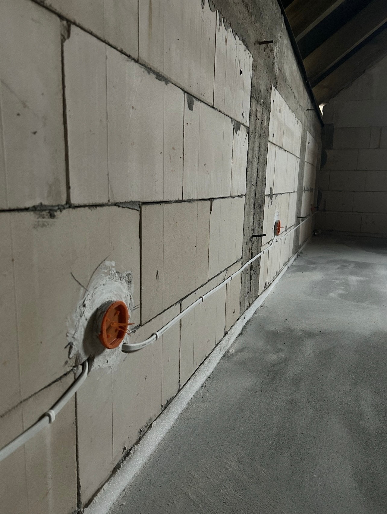 Wewnętrzna instalacja elektryczna w surowym budynku: białe kable prowadzone wzdłuż ściany z pustaków, pomarańczowe puszki pod gniazdka. Szara podłoga.