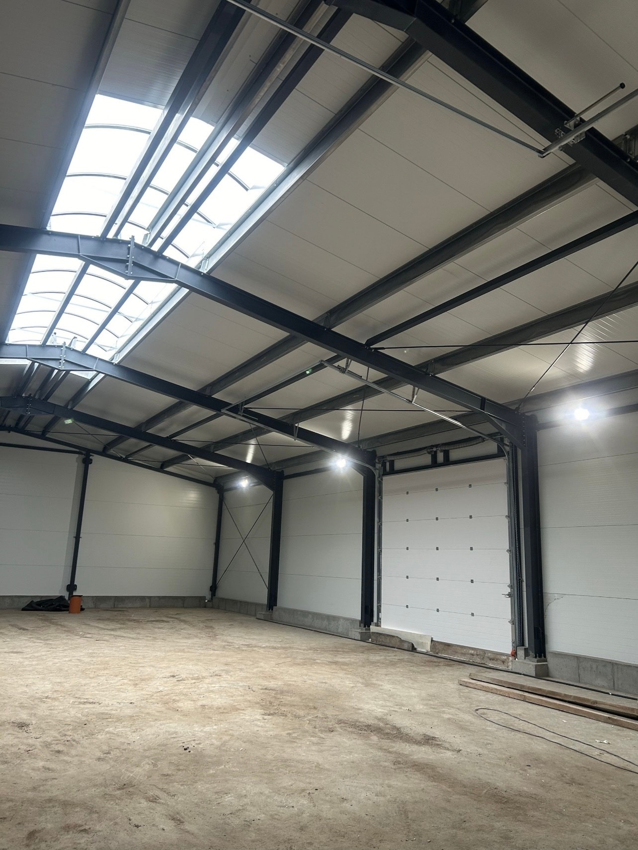 Wnętrze hali magazynowej z oświetleniem LED, widoczne stalowe konstrukcje, białe ściany z paneli i brama segmentowa. Na suficie świetliki doświetlające wnętrze.