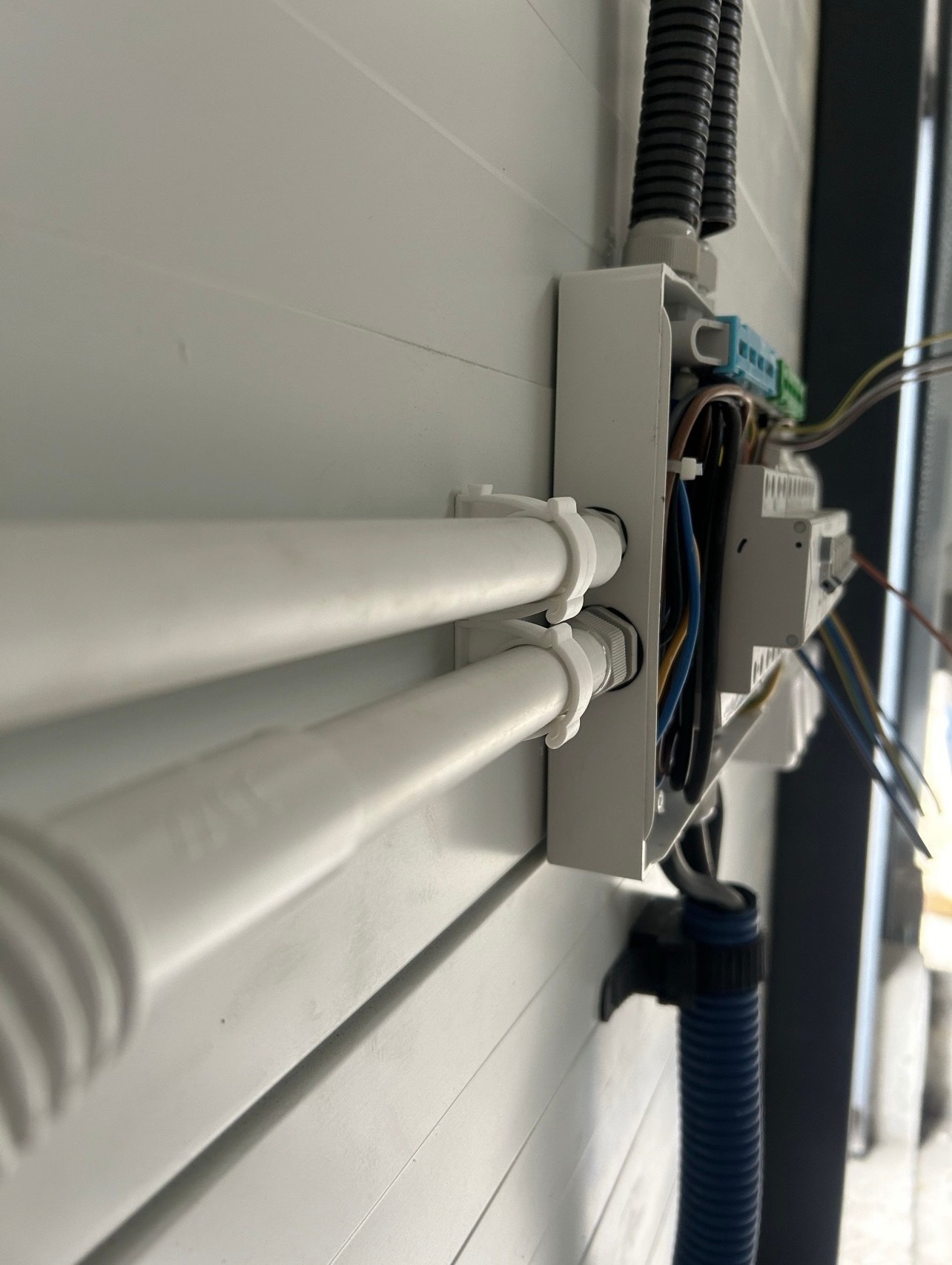 Biała puszka instalacyjna z przewodami i rurkami ochronnymi przymocowana do ściany o panelowej strukturze. Widoczne złącza i kolorowe kable.
